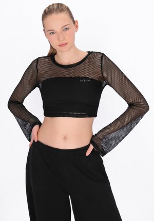 Junge Frau mit blondem Haar trägt ein schwarzes, langärmeliges Netz-Crop-Top über einem schwarzen Bandeau, kombiniert mit lockeren schwarzen Hosen, posiert mit den Händen in die Hüften gestützt.