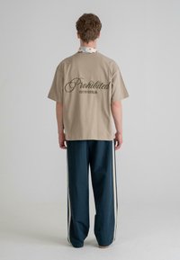 Beige pamut póló "Tiltva" és "10119 BERLIN" felirattal a hátsó részen; sötétkék, széles lábú nadrággal párosítva, fehér oldalsávokkal.