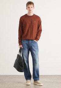 Maglione a maglia marrone, jeans blu e sneakers beige. Modello che tiene una borsa tote nera. Look semplice e rilassato.