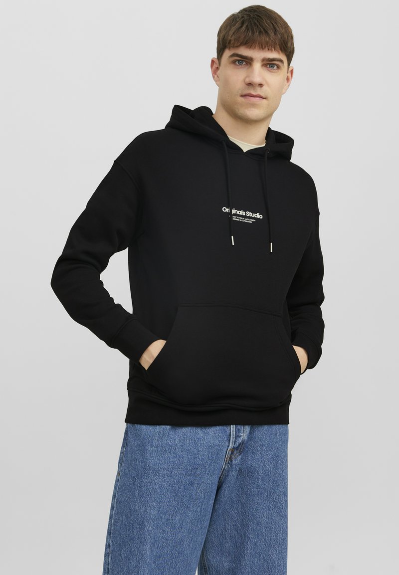 Zwarte hoodie met trekkoord, voorzak en witte tekstlogo. Voorzien van ribbelboorden en onderzoom. Gecombineerd met lichtblauwe jeans.