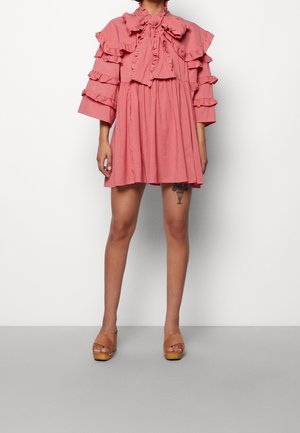 Różowa sukienka z falbankami, z wysokim kołnierzem i rozszerzanymi rękawami, z dopasowaną talią i rozkloszowanym dołem. Do tego brązowe sandały na koturnie.