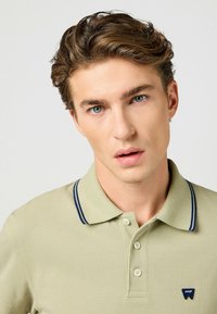 Kaki pikétröja med texturerad yta, krage med blå ränder, knäppning med tre knappar och liten blå logotyp på bröstet.