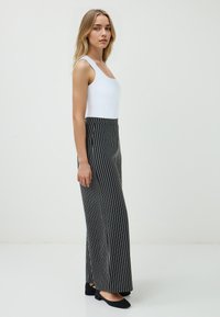 Een jonge vrouw staat zijwaarts, gekleed in een witte tanktop en zwart-wit gestreepte brede broek, tegen een effen achtergrond.