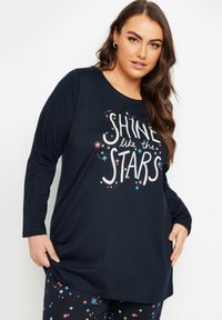 Långärmad marinblå topp med texten "SKINA som STJÄRNORNA" i vitt och färgglada stjärndekorationer. Tillsammans med svarta byxor prydda med stjärnmönster.