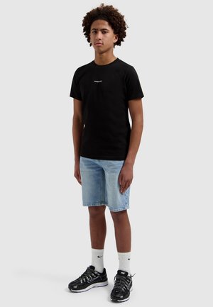 Jong persoon met krullend haar, een zwart t-shirt, licht denim short, witte Nike sokken en zwarte Nike sneakers op een lichte achtergrond.