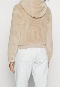 Beżowy polarowy hoodie o miękkiej fakturze, z kapturem na sznurku i prążkowanym dołem, zestawiony z jasnoniebieskimi spodniami.