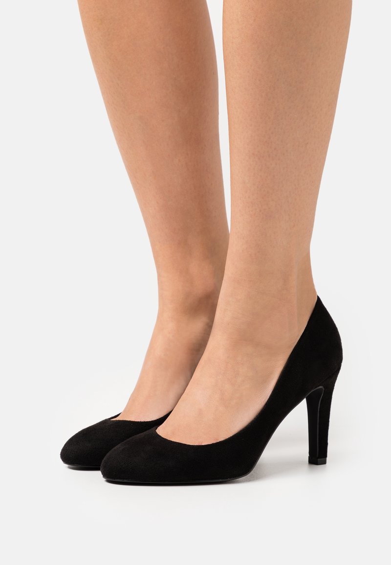 Femme portant des escarpins en daim noir à talons hauts, jambes visibles à partir du milieu du mollet, sur fond blanc.