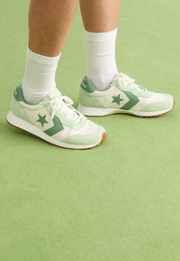 Grønne sneakers med hvite detaljer, laget av semsket skinn og syntetiske materialer, med en stjerne-logo og hvite snørebånd, satt mot en myk grønn bakgrunn.