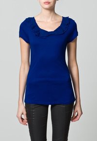 Blå kortärmad t-shirt med volangkrage, figurnära silhuett och mjuk textur. Matchas med svarta slim fit-byxor.