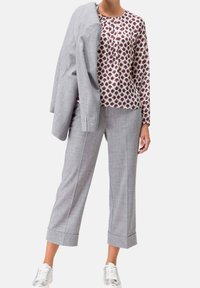 Femme portant un pantalon gris court, un blouse à manches longues à motifs, des baskets blanches, tenant un blazer gris assorti sur une épaule.