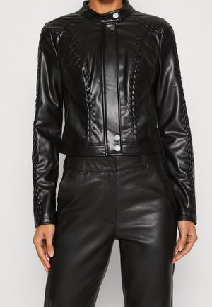 Nainen, joka pukeutuneena mustaan keinonahkatakkiin, jossa on etuosan painonapit, ja vastaaviin housuihin, kädet rennosti sivuilla neutraalia taustaa vasten.