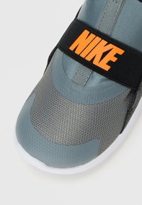 Sneaker Nike grigio con texturizzazione a rete sul davanti, dotato di una fascia elastica nera con il logo Nike arancione che attraversa la parte superiore.