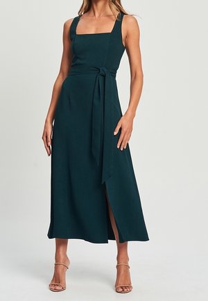 Woman wearing a sleeveless dark green midi dress with a square neckline, waist tie, side slit, and beige high-heeled sandals.