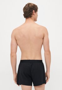 Male model seen from back, shirtless, wearing black boxer shorts against plain white background.