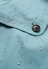 Wodoodporny materiał w kolorze turkusowym z kroplami wody, ciemny guzik oraz widoczne szwy. Gładka tekstura.