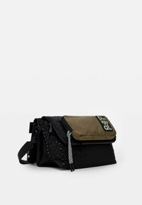 Black woven fabric bag with an olive green flap. Features multiple zippered pockets and a patterned strap. Branding displayed on the flap.