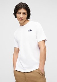 Νέος άνδρας με μεσαίου μήκους σκούρα μαλλιά φοράει λευκό μπλουζάκι The North Face και μπεζ παντελόνια με τα χέρια στις τσέπες, μπροστά σε απλό φόντο.