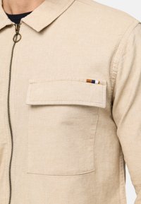 Veste beige avec une poche à rabat devant et une fermeture éclair, portée sur une chemise noire, montrant les détails du haut de la poitrine et des épaules.