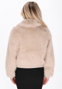 Person med langt lyst hår iført en beige kunstpelsjakke og sort nederdel, vist fra ryggen mod en hvid baggrund.