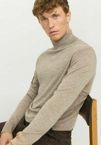 Beżowy sweter z wysokim kołnierzem, wykonany z miękkiej, drobno dzianinowej tkaniny, z długimi rękawami i dopasowanym krojem. Noszony na ciemne spodnie.