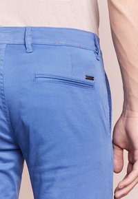 Ljusblå byxor med en jämn textur, utrustade med en bakficka och en liten metallaccent på sidan av fickan.