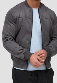 Veste bomber en daim gris avec poignets et col côtelés, comprenant une fermeture éclair et deux poches avant. Portée par-dessus une chemise texturée bleu clair.