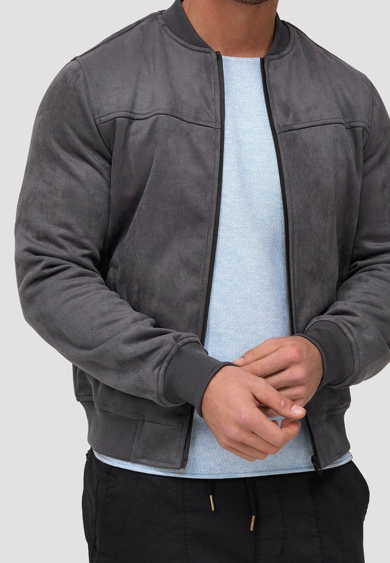 Veste bomber en daim gris avec poignets et col côtelés, comprenant une fermeture éclair et deux poches avant. Portée par-dessus une chemise texturée bleu clair.