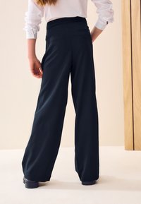 Pantaloni a gamba larga in tessuto blu navy scuro. Presentano una vita alta con tasche posteriori. Texture liscia e design minimalista. Scarpe nere sono visibili in fondo.