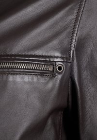 Nahaufnahme einer braunen Lederjacke mit einer Reißverschlusstasche, glatter Textur und sichtbaren Nähten entlang der Kanten. Akzentuiert mit silbernem Metallzubehör.