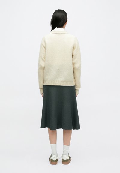 Μπεζ πουλόβερ από μάλλινο ύφασμα με ψηλό γιακά, συνδυασμένο με σκούρα πράσ�ινη φούστα. Λευκές κάλτσες και ελιά-πράσινα παπούτσια ολοκληρώνουν την εμφάνιση.