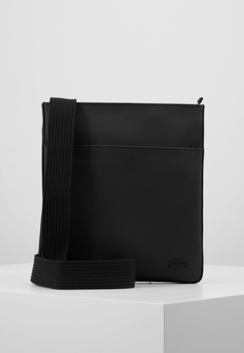 Sac bandoulière noir avec une large sangle texturée, design simple et petit logo embossé dans le coin inférieur droit, présenté sur une surface blanche.