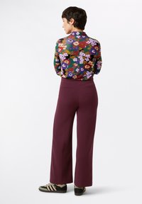 Camicia a maniche lunghe con motivo floreale; pantaloni larghi bordeaux; sneakers nere e verdi. Colori e texture vivaci visibili.