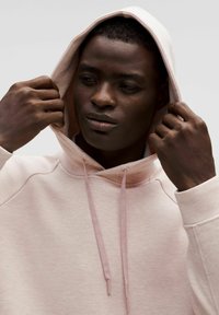 Sudadera rosa claro con cordón, que presenta un dobladillo y puños acanalados. El modelo ajusta la capucha, mostrando una textura suave y un ajuste relajado.
