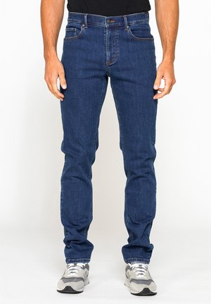 Uomo che indossa jeans slim fit blu e sneakers grigie, in piedi con le mani rilassate lungo i fianchi su sfondo bianco.