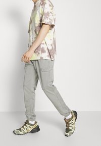 Persona che cammina indossando una maglietta tie-dye, pantaloni cargo grigi chiari con polsini elasticizzati, calze bianche e scarpe da trail in verde e beige.