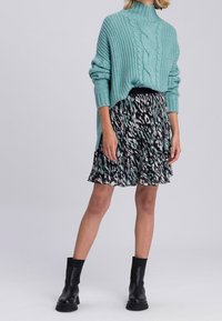Pull en tricot turquoise à col montant, associé à une jupe à volants avec un motif noir et blanc. Le modèle porte des bottines noires.