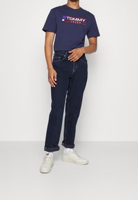 Mörkblå bomulls t-shirt med "TOMMY JEANS"-logotyp, i kombination med mörka denimjeans och vita sneakers. Jeansen har uppkavlade ärmar.