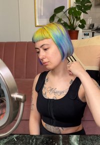 Kort svart crop top med axelband; flervägsfärgat hår (blått, grönt, gult); tatueringar synliga på armar och bröst; rund spegel i förgrunden.