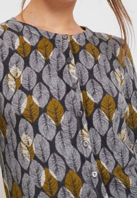 Cardigan mit einem Blättermuster in Grau, Gelb und Weiß. Verfügt über einen runden Ausschnitt und fünf graue Knöpfe vorne. Strukturierter Stoff.