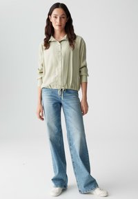 Lichtgroene, langarmige blouse met knoopsluiting en een trekkoord onderaan, gecombineerd met hoogzwaaiende lichtblauwe wijde jeans en witte sneakers.