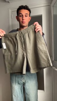 Jeune homme tenant un short beige avec une taille élastique et un cordon dans une pièce avec une porte blanche et un miroir.