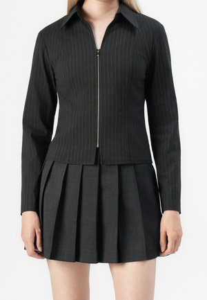 Femme portant une veste noire à fines rayures avec fermeture éclair et manches longues, associée à une jupe plissée foncée sur un fond uni.