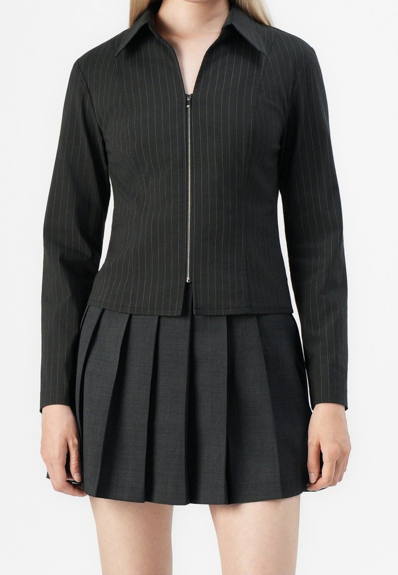 Femme portant une veste noire à fines rayures avec fermeture éclair et manches longues, associée à une jupe plissée foncée sur un fond uni.