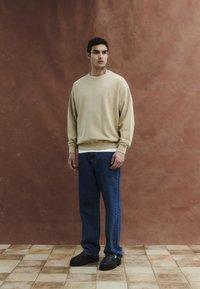 Férfi áll barna, texturált fal előtt, bézs pulóvert, kék farmert és fekete cipőt visel a csempézett padlón, enyhén jobbra néz.