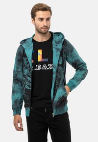 Kapuzensweatjacke in Teal mit schwarzen Spritzmustern; ausgestattet mit einer Fronttasche und gerippten Bündchen, getragen über einem schwarzen Grafik-T-Shirt.