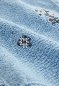 Tissu en denim bleu clair avec des détails de broderie florale comprenant des pétales blancs, un centre rouge et des feuilles vertes. Texture douce visible.