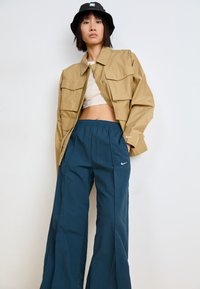 Χακί κοντό μπουφάν με μεγάλες τσέπες, συνδυασμένο με φαρδιά τιρκουάζ παντελόνια Nike με ελαστική μέση και πλαϊνές τσέπες. Μαύρο καπέλο bucket.