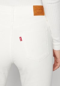 Primer plano del bolsillo trasero de unos jeans Levi's blancos con etiqueta roja de Levi's y parche de cuero marrón de Levi Strauss & Co. en la cinturilla, combinados con una camiseta blanca de canalé.