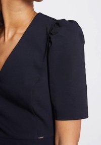 Navyblaues Kleid mit V-Ausschnitt, kurzen Puffärmeln und glatter Textur. Bemerkenswertes Detail: kleines Hardware-Accessoire in der Nähe des Saums.