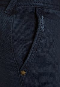 Mörkblå denimbyxor med en framficka som har en mässingsknapp och synlig sömnad, som visar en strukturerad tygkvalitet.